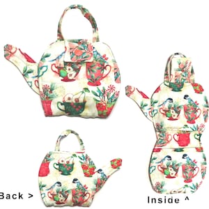 May include: A fabric teapot-shaped tea cozy with a handle and spout. The cozy is cream-colored and covered in a pattern of red teacups, pears, and birds. The back of the cozy is visible, along with the inside, showing the same pattern.