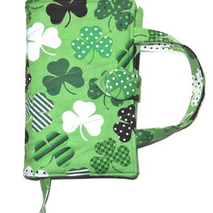 May include: A green quilted book cover with a handle, featuring a pattern of shamrocks in various shades of green, white, and black. The cover has a button closure and a bookmark ribbon.