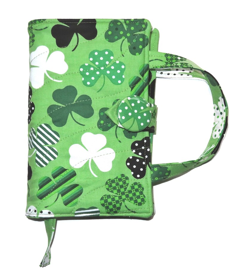 May include: A green quilted book cover with a handle, featuring a pattern of shamrocks in various shades of green, white, and black. The cover has a button closure and a bookmark ribbon.