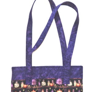 May include: A fabric tote bag featuring a dark pattern of shelves filled with potion bottles, skulls, and pumpkins. The bag has a solid purple mottled fabric border at the top and two matching purple fabric shoulder straps extending upwards.