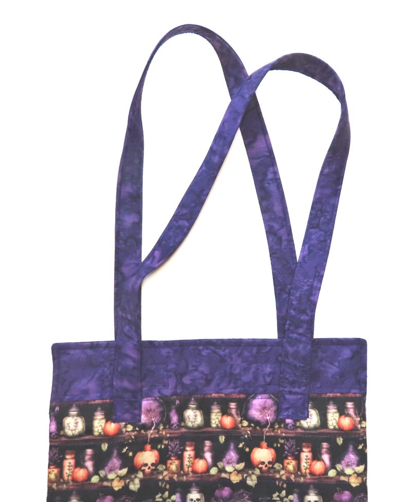 May include: A fabric tote bag featuring a dark pattern of shelves filled with potion bottles, skulls, and pumpkins. The bag has a solid purple mottled fabric border at the top and two matching purple fabric shoulder straps extending upwards.