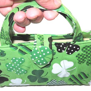 May include: A green fabric handbag with a shamrock pattern. The bag features a handle, a button closure, and a variety of shamrock designs in white, black, and green. The bag is held by a hand.