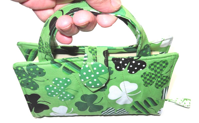 May include: A green fabric handbag with a shamrock pattern. The bag features a handle, a button closure, and a variety of shamrock designs in white, black, and green. The bag is held by a hand.