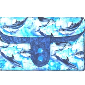 May include: A fabric wallet with a blue and white dolphin pattern. The wallet has a flap closure with a dark blue accent and two light blue buttons. The background has a watercolor effect.