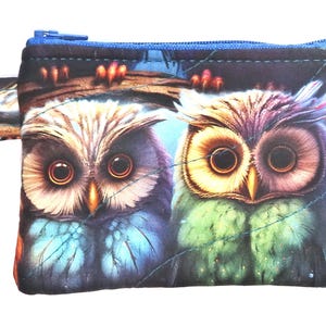May include: A small, rectangular coin purse featuring a vibrant owl design. The purse has a blue zipper and a key ring attachment. The owl illustration showcases detailed feathers in shades of blue, green, and purple, set against a dark background.