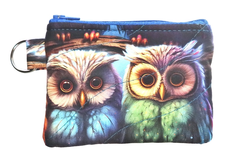 May include: A small, rectangular coin purse featuring a vibrant owl design. The purse has a blue zipper and a key ring attachment. The owl illustration showcases detailed feathers in shades of blue, green, and purple, set against a dark background.