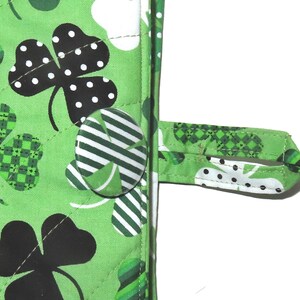 May include: Close-up of a green quilted fabric with a St. Patrick's Day theme. The fabric features large black shamrocks with white polka dots, smaller green shamrocks, and a button closure. A fabric loop is attached for hanging.
