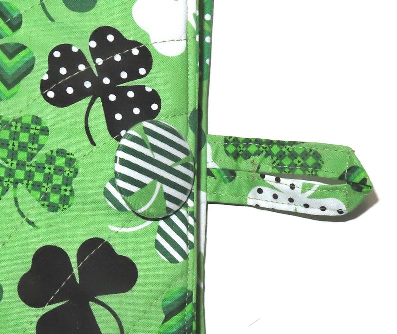 May include: Close-up of a green quilted fabric with a St. Patrick's Day theme. The fabric features large black shamrocks with white polka dots, smaller green shamrocks, and a button closure. A fabric loop is attached for hanging.