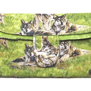 May include: A quilted clutch featuring a repeating pattern of wolves in a grassy field. The clutch has a green flap with a snap closure. The wolves are depicted in various poses, with some lying down and others standing.