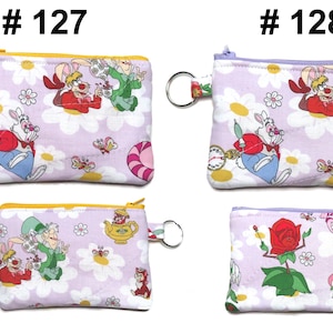 May include: Four small, rectangular coin purses with zipper closures and key rings. The purses feature an Alice in Wonderland theme with characters like the White Rabbit and Cheshire Cat on a light purple background with white daisies.