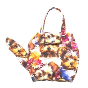 Pode incluir: Uma bolsa de mão única em forma de bule, com um padrão repetido de filhotes de Yorkshire Terrier e detalhes florais. A bolsa tem uma alça e um pequeno fecho de aba, com uma paleta de cores vibrante de marrom, branco, rosa e amarelo.