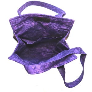 May include: A quilted tote bag featuring a vibrant purple batik fabric with a mottled pattern. The bag has an open top, two fabric shoulder straps, and an interior lining with a horizontal pocket along the back wall of the bag's main compartment.
