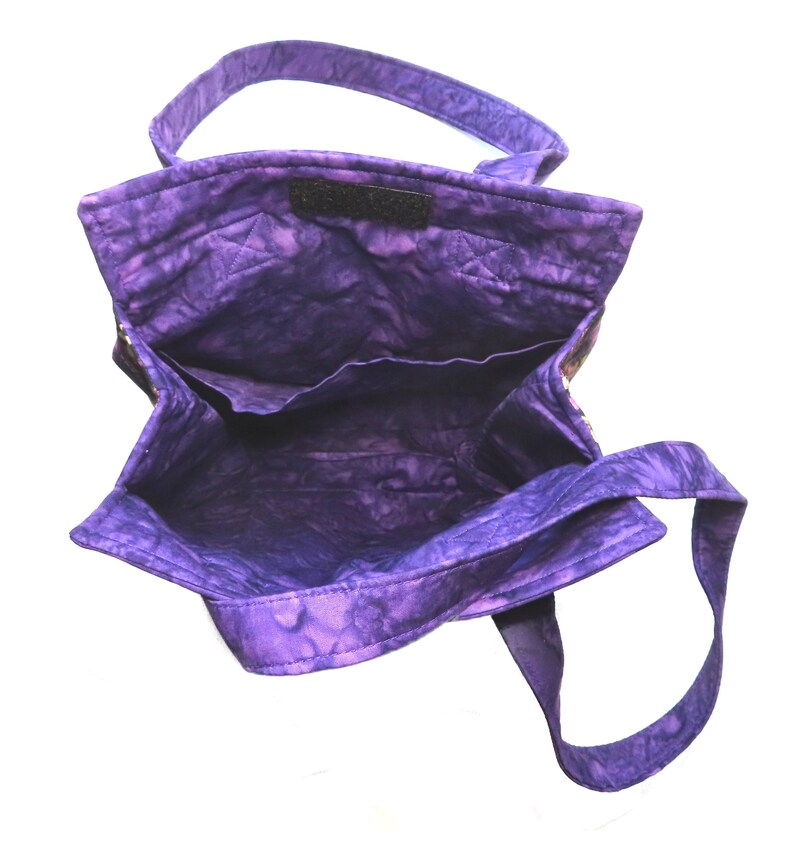 May include: A quilted tote bag featuring a vibrant purple batik fabric with a mottled pattern. The bag has an open top, two fabric shoulder straps, and an interior lining with a horizontal pocket along the back wall of the bag's main compartment.