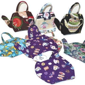 May include: A collection of teapot-shaped fabric tea cozies in various patterns. Designs include floral, fruit, and patterned prints. One cozy features a label that reads "Black Tea". Each cozy has a handle and spout, designed to fit over a teapot.