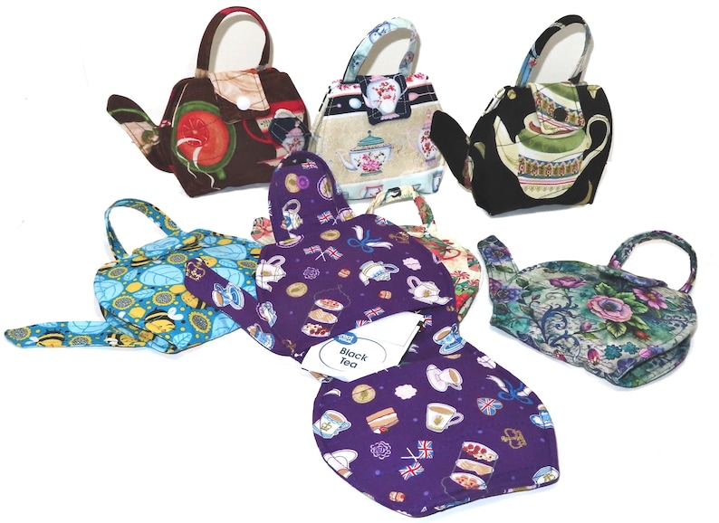 May include: A collection of teapot-shaped fabric tea cozies in various patterns. Designs include floral, fruit, and patterned prints. One cozy features a label that reads "Black Tea". Each cozy has a handle and spout, designed to fit over a teapot.