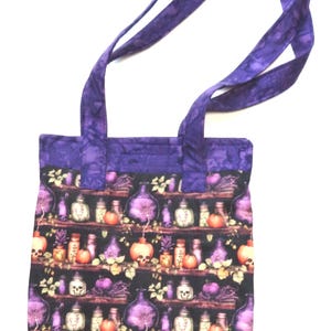 May include: A fabric tote bag featuring a repeating pattern of shelves filled with potion bottles, skulls, and pumpkins on a black background. The bag has a solid purple batik-style fabric trim at the top and two matching purple fabric shoulder straps.