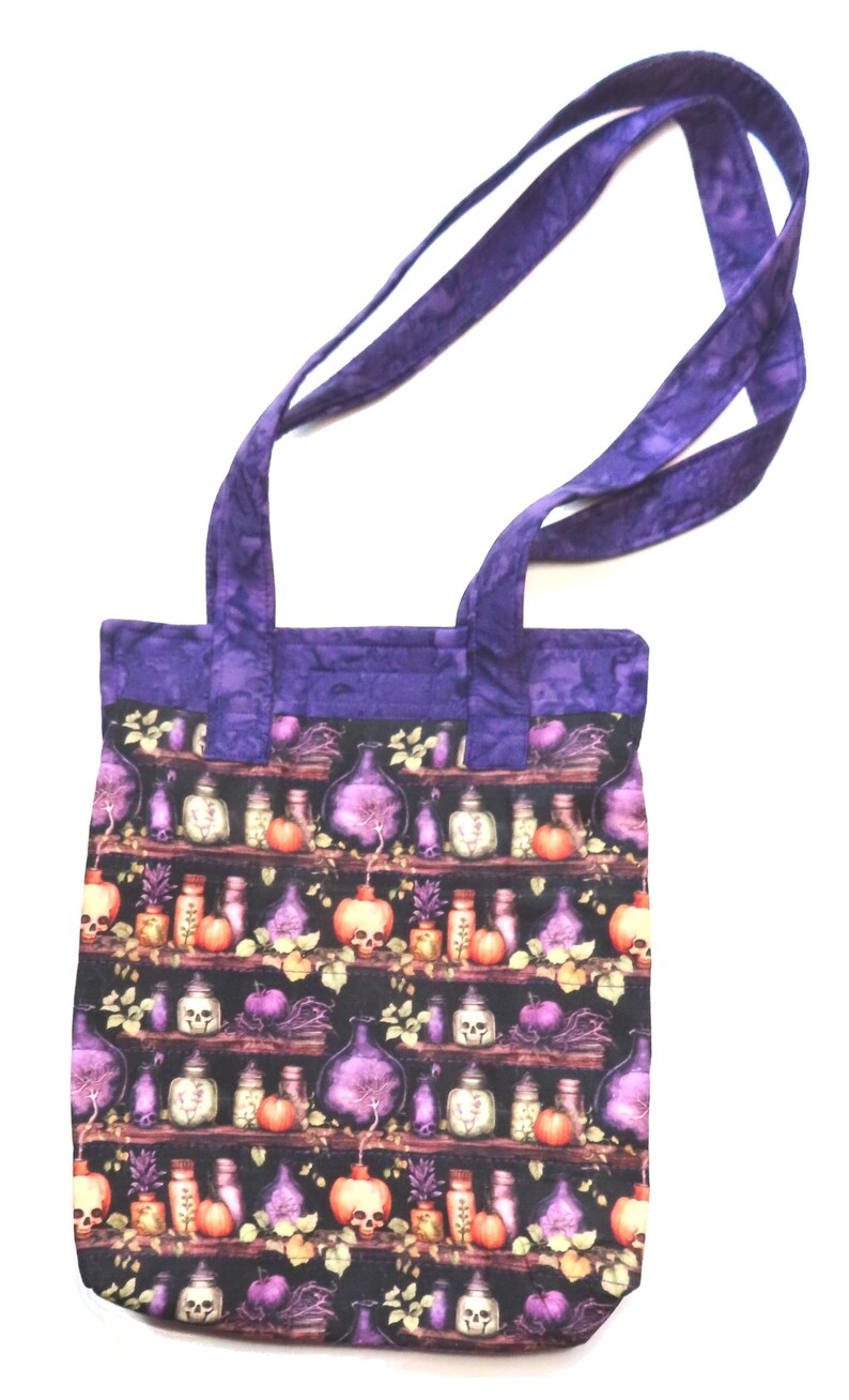 May include: A fabric tote bag featuring a repeating pattern of shelves filled with potion bottles, skulls, and pumpkins on a black background. The bag has a solid purple batik-style fabric trim at the top and two matching purple fabric shoulder straps.