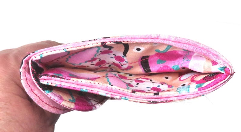 May include: A folded, pink fabric wallet with a colorful interior. The interior features a pattern of pink, peach, and teal abstract shapes. The exterior is a solid pink with a black border.