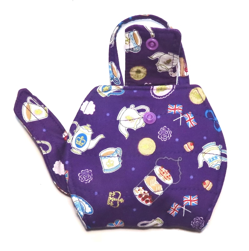 May include: A purple tea cozy with a teapot shape. The cozy is covered in a pattern of teacups, teapots, and British flags. The handle is white. The cozy is designed to keep tea warm.