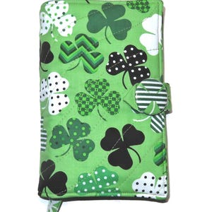 May include: A green quilted fabric cover with a repeating pattern of shamrocks in various designs, including polka dots and checkered patterns. The cover has a button closure and a small tab. The shamrocks are in black, white, and shades of green.