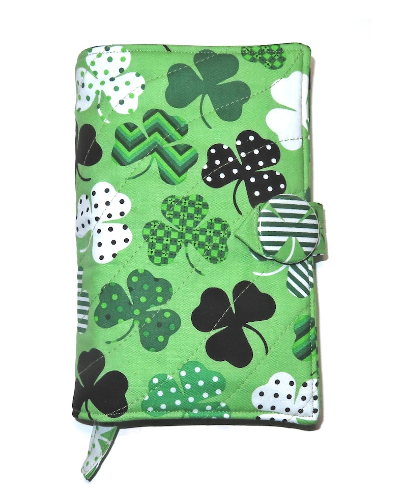 May include: A green quilted fabric cover with a repeating pattern of shamrocks in various designs, including polka dots and checkered patterns. The cover has a button closure and a small tab. The shamrocks are in black, white, and shades of green.