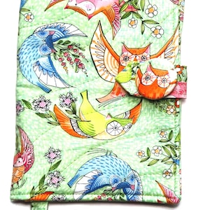 May include: A quilted journal cover with a light green background and a repeating pattern of colourful owls, birds, and floral elements. The cover has a button closure and a matching bookmark. The design features orange, blue, pink, and yellow hues.