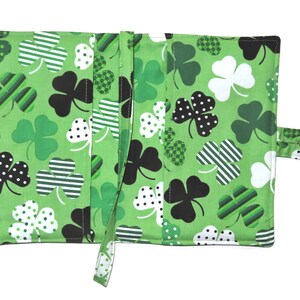 May include: Green fabric wallet featuring a repeating pattern of shamrocks in various designs: black polka dots, white polka dots, and striped. The wallet has a strap closure and is open, revealing interior compartments.