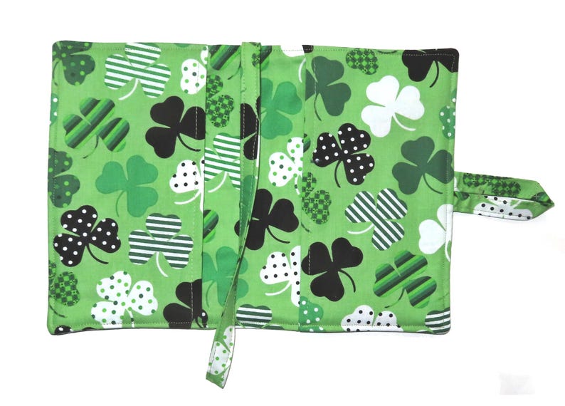 May include: Green fabric wallet featuring a repeating pattern of shamrocks in various designs: black polka dots, white polka dots, and striped. The wallet has a strap closure and is open, revealing interior compartments.