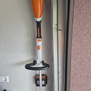 May include: An orange and white cordless string trimmer with a black handle and a black loop handle. Two black battery packs are attached to the trimmer's shaft. The trimmer is mounted on a wall.