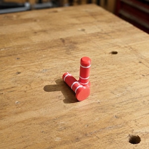 May include: Two cylindrical red pegs with flat circular bases and two thin white horizontal stripes encircling the upper portion of each shaft. The objects are positioned upright and angled on a flat surface, displaying a smooth matte finish.
