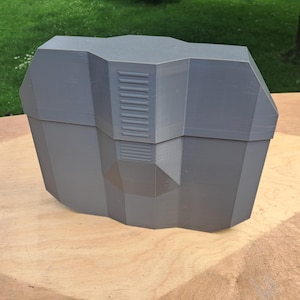 Op de afbeelding: Een matte grijze, 3D-geprinte opbergdoos met een geometrisch, hoekig ontwerp. De doos heeft een veelzijdige buitenkant met een centraal verticaal paneel met horizontale geribbelde details, wat zorgt voor een gestructureerde, industriële uitstraling.