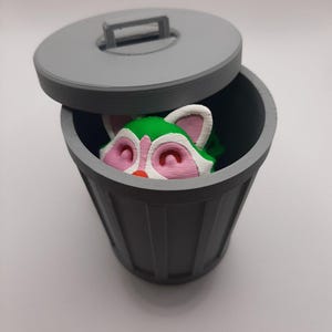 Puede incluir: Un cubo de basura de plástico gris con una tapa que está parcialmente abierta. Una pequeña figura de mapache verde y rosa está dentro del cubo de basura.