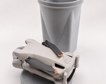 Könnte beinhalten: Eine hellgraue faltbare Drohne mit schwarzen Propellern mit orangefarbenen Spitzen, die auf einer runden grauen Basis neben einem passenden zylindrischen grauen Aufbewahrungsbehälter mit Gewinderand und Trennwand steht.