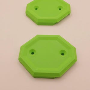 Puede incluir: Dos objetos verdes brillantes, de forma octogonal, cada uno con dos pequeños agujeros. Los objetos están apilados verticalmente sobre un fondo claro. Los objetos parecen ser de plástico.