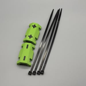 Op de afbeelding: Een set van vier zwarte kabelbinders en twee limoengroene plastic componenten met kruisvormige en rechthoekige uitsparingen. De items zijn gerangschikt op een lichtgrijze ondergrond.