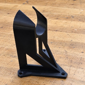 May include: A black 3D-printed wall mount bracket featuring a curved cradle top for holding cylindrical objects. The structure has a triangular support frame with a flat base plate containing two circular screw holes for secure mounting.
