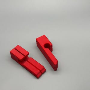 May include: Two bright red wooden clothespins are displayed against a light gray background. One pin is lying flat, while the other stands upright. The pins have a classic design with a spring mechanism.