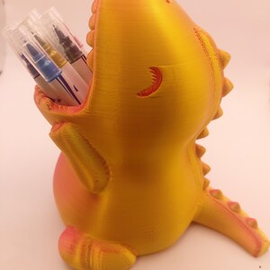 May include: A 3D-printed, yellow and orange dinosaur-shaped pen holder. The dinosaur has an open mouth to hold pens and markers. The holder is designed with a textured surface and a small tail.