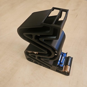 Puede incluir: Un soporte de baterías de plástico negro impreso en 3D con dos baterías AA en su interior. Las baterías están etiquetadas con "Duracell" y "Energizer".