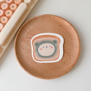 May include: A sticker featuring a cartoon bear face inside a slice of toast. The bear has a light green face with a brown nose and black eyes. The toast is brown with a light orange inside. The sticker is on a wooden surface.