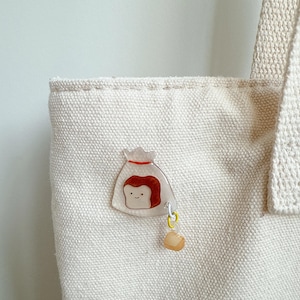 May include: A clear acrylic pin featuring a cartoon illustration of a bag with a face and a small charm. The pin is attached to a cream-colored canvas tote bag. The bag has a simple design and is made of a sturdy material.