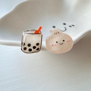 May include: Two small resin charms, one shaped like a boba tea with a red straw and black boba pearls, and the other shaped like a white dumpling with a smiling face.