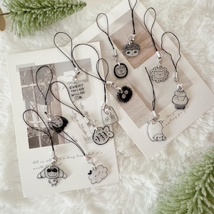May include: A collection of black and white cartoon character phone charms. Each charm is attached to a black lanyard with a silver clasp. The charms feature various designs, including a bear, a cloud, and a character with the text "EVERY THING WILL BE OK".