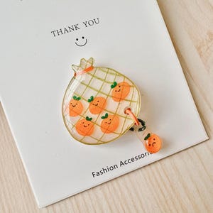 May include: A clear plastic charm with a checkered pattern and a design of orange slices with smiley faces. The charm is attached to a smaller orange slice charm with a sad face. The text "THANK YOU" and a smiley face are visible on a white background.