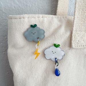 May include: Two cute cloud-shaped charms with a lightning bolt and a raindrop hanging from them. The charms are made of white and gray plastic and have green leaves on top. The charms are attached to a white canvas bag.