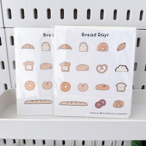 May include: Two packs of stickers featuring various bread illustrations, including croissants, toast, and bagels. The stickers are in a clear plastic sleeve, with the words "Bread Days" printed on the front. The stickers are in shades of brown and beige.