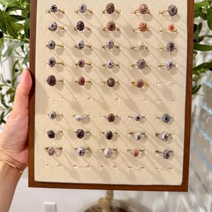 May include: A display of gold-toned rings with various gemstone settings. The rings are arranged on a beige surface within a wooden frame. The gemstones feature a range of colors, including shades of purple, brown, and silver.