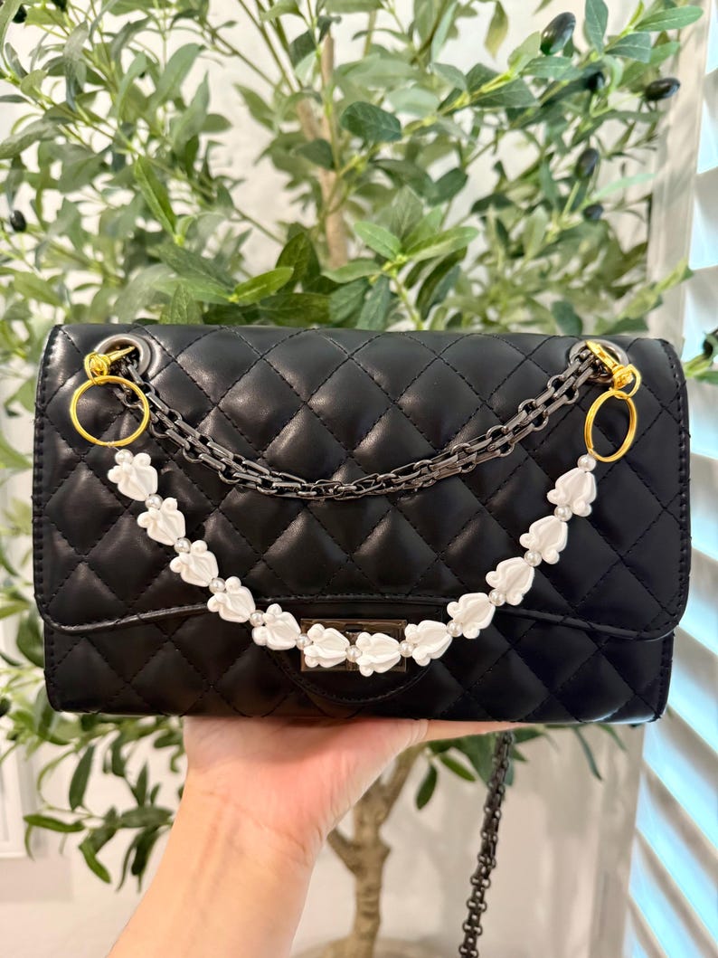 Pu&ograve; includere: Borsa nera trapuntata con tracolla a catena e dettagli floreali bianchi. La borsa presenta finiture color oro e chiusura con patta. La tracolla &egrave; una combinazione di una catena di metallo e una catena di perline bianche.