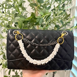Pu&ograve; includere: Borsa trapuntata nera con manico floreale bianco e tracolla a catena argentata. La borsa presenta finiture color oro e una forma rettangolare. Lo sfondo include fogliame verde.
