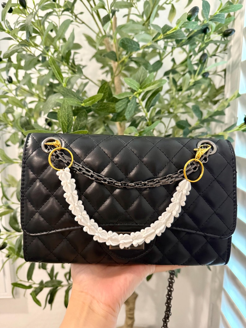 Pu&ograve; includere: Borsa trapuntata nera con manico floreale bianco e tracolla a catena argentata. La borsa presenta finiture color oro e una forma rettangolare. Lo sfondo include fogliame verde.